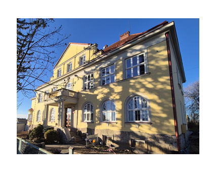 Zabytkowy, dwupiętrowy budynek o ż&oacute;łtej elewacji z białymi oknami i czerwonym dachem, widziany z zewnątrz w słoneczny dzień. Przed budynkiem drzewa rzucają cienie na fasadę.