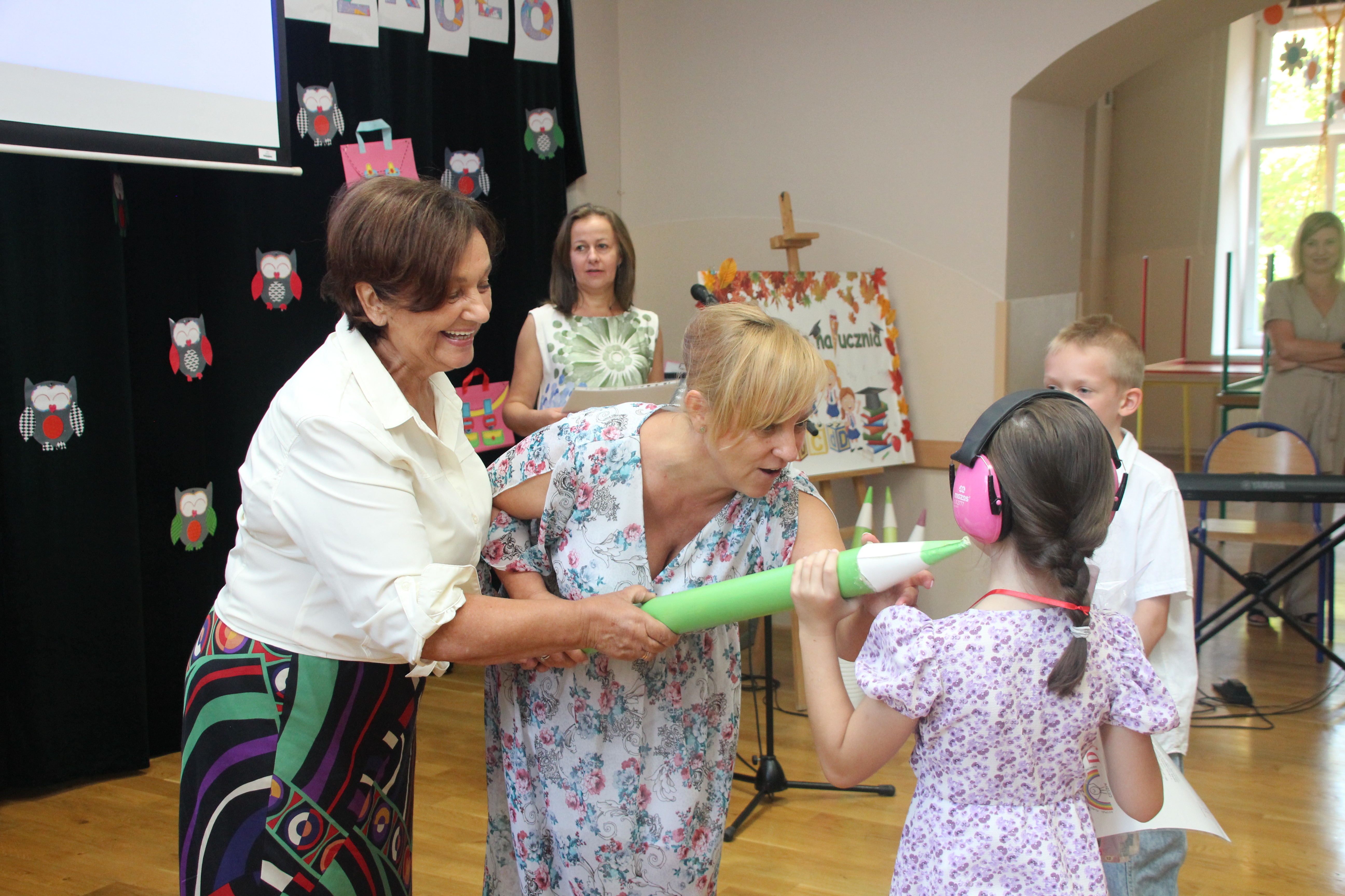Janina Żagan Starosta Cieszyński i Dyrektor szkoły uśmiechnięte trzymają duży zielony oł&oacute;wek, kt&oacute;rym dotykają dziewczynkę w r&oacute;żowej sukience i ochronnikach słuchu, uczestniczącą w szkolnej uroczystości.