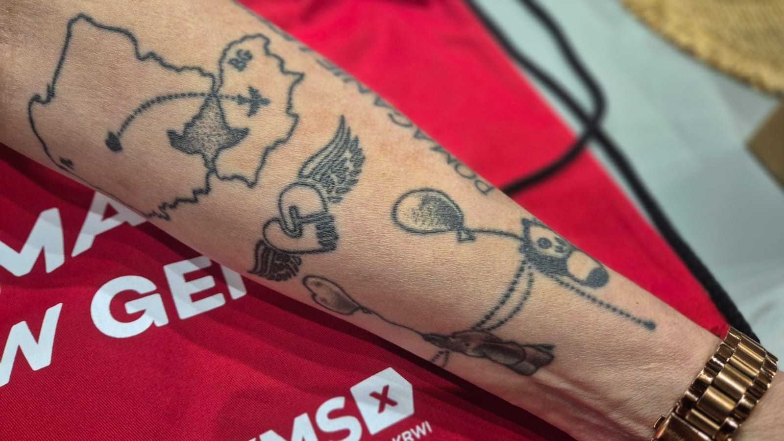 Przedramię z tatuażem przedstawiającym mapę Polski z trasą lotu, serce ze skrzydłami oraz balonikowego pieska; tło stanowi czerwona koszulka z logo Fundacji DKMS, widoczny fragment złotego zegarka na nadgarstku.
