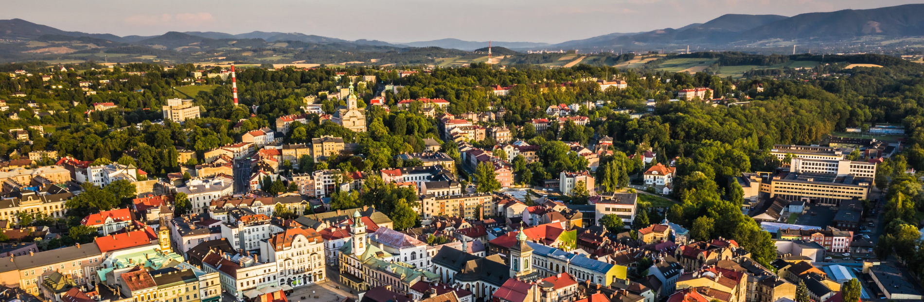 Widok miasta Cieszyn otoczonego zielenią i wzgórzami. W centrum historyczne budynki z czerwonymi dachami, wśród nich ratusz z wieżą. W tle góry i tereny wiejskie. Przestrzeń pełna drzew i parków, harmonijnie łącząca architekturę z naturą.