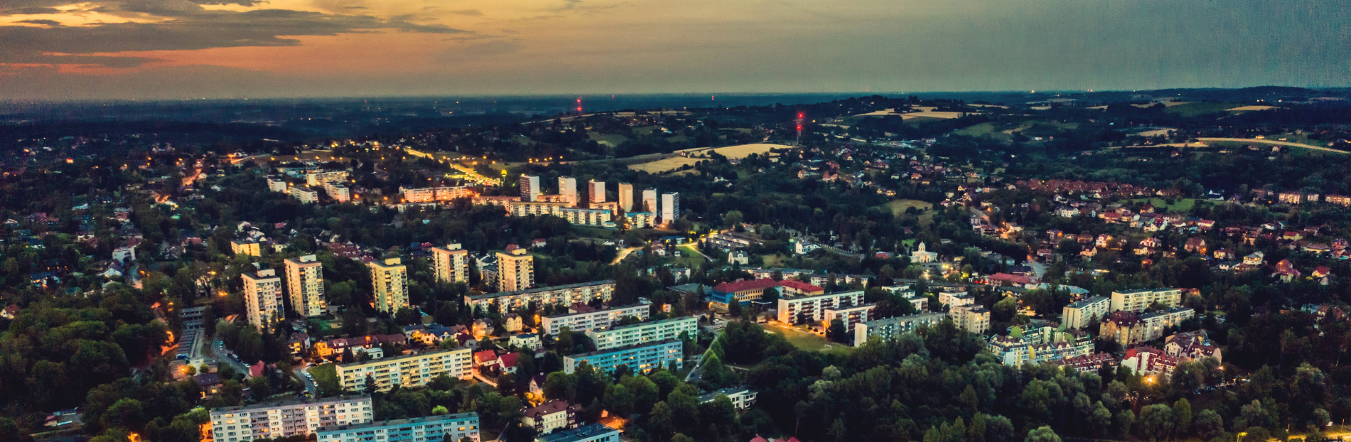 Rozległa panorama miasta Ustroń o zmierzchu, ukazująca układ urbanistyczny z blokami mieszkalnymi i domami jednorodzinnymi. Ulice rozświetlone są światłami latarni, tworząc kontrast z ciemniejącym niebem. W tle rozciągają się pola i pagórkowaty krajobraz.