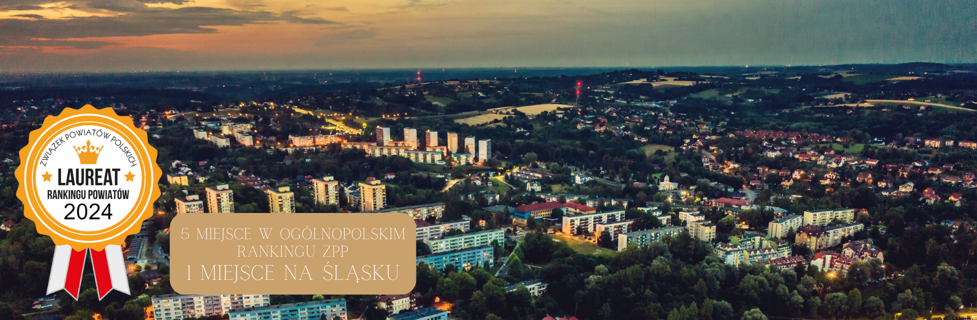 Rozległa panorama miasta Ustroń o zmierzchu, ukazująca układ urbanistyczny z blokami mieszkalnymi i domami jednorodzinnymi. Ulice rozświetlone są światłami latarni, tworząc kontrast z ciemniejącym niebem. W tle rozciągają się pola i pagórkowaty krajobraz.