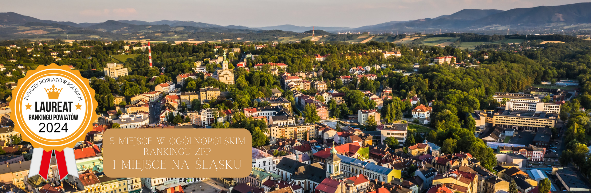 Widok miasta Cieszyn otoczonego zielenią i wzgórzami. W centrum historyczne budynki z czerwonymi dachami, wśród nich ratusz z wieżą. W tle góry i tereny wiejskie. Przestrzeń pełna drzew i parków, harmonijnie łącząca architekturę z naturą. W lewym rogu zło