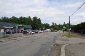 Ulica z popękaną nawierzchnią prowadzi obok sklepu i przystanku autobusowego po lewej stronie. Po prawej widoczny jest chodnik, ogrodzenie i znak przejścia dla pieszych. W tle rosną drzewa i poruszają się pojazdy.