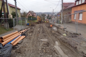 Rozkopana ulica z błotnistym podłożem, otoczona barierkami ochronnymi. Po lewej stronie leżą rury kanalizacyjne, a w oddali widoczna jest koparka. Po prawej mijana jest przychodnia stomatologiczna w pomarańczowym budynku.