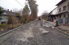 Ulica w trakcie przebudowy – nawierzchnia pokryta tłuczniem, z wytyczonymi krawężnikami. Po bokach stoją znaki ostrzegawcze, a w oddali widoczna jest koparka i kilku pracowników. Domy po obu stronach drogi.