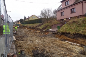 Droga w trakcie głębokiej przebudowy – błotnista nawierzchnia, wykop i widoczne rury. Po lewej stronie pracownicy w kamizelkach odblaskowych, dalej koparka. Domy jednorodzinne stoją przy skarpie z trawnikiem.