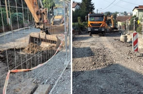 Widok rozkopanej ulicy z ciężkimi maszynami budowlanymi – koparką i wywrotką. Droga jest zamknięta metalowym ogrodzeniem, a nawierzchnia zerwana do warstwy ziemi i tłucznia. Trwają prace ziemne.