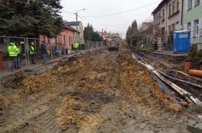 Rozkopana ulica z głębokim wykopem biegnącym przez środek jezdni, po bokach ułożone rury i przewody. Kilku pracowników w odblaskowych kurtkach stoi przy ogrodzeniu, a w tle widoczny sprzęt budowlany.
