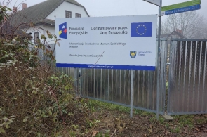 Metalowe ogrodzenie z zamontowaną tablicą informacyjną o dofinansowaniu z Unii Europejskiej. W tle budynek muzeum, krzewy i drzewa bez liści. Po prawej stronie widoczny fragment bramy i znak drogowy.