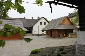 Enklawa - Chata kumornika i kuźnia. Dziedziniec z drewnianymi budynkami o dachach pokrytych gontem; w tle biały budynek murowany, na pierwszym planie gałęzie drzewa.