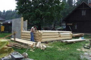 Enklawa w trakcie budowy. Budowa drewnianego domu z bali; na pierwszym planie mężczyzna pracujący przy konstrukcji, w tle inne drewniane budynki i drzewa.