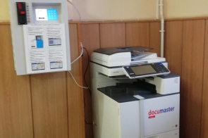 Urządzenie wielofunkcyjne do druku i kopiowania oraz panel systemu documaster zamontowany na ścianie obok.