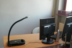 Stanowisko komputerowe z dwoma monitorami, klawiaturą, dokumentami oraz kamerą dokumentacyjną ustawioną na biurku.