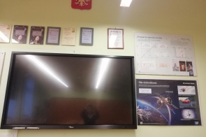 Monitor interaktywny zamontowany na ścianie, powyżej widoczne godło Polski i dyplomy, obok plansze edukacyjne.