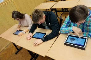 Trzech uczniów siedzi w klasie przy ławkach i pracuje na tabletach, na ekranach widoczne modele trójwymiarowe.