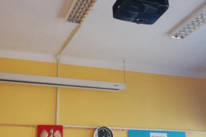 Projektor zamontowany pod sufitem w sali lekcyjnej, poniżej godło Polski, zegar ścienny i plansza edukacyjna.