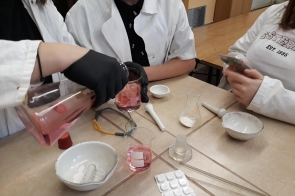 Grupa uczniów w fartuchach laboratoryjnych przy stole, jedna osoba przelewa różowy płyn z cylindry do zlewki, wokół leżą naczynia i przybory chemiczne.