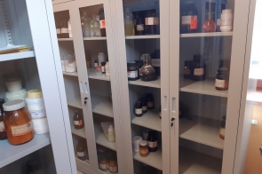 Metalowe szafy z przeszklonymi drzwiami, wypełnione pojemnikami i butelkami z odczynnikami chemicznymi, ustawione w pomieszczeniu pracowni.