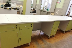 Długi blat roboczy pokryty białymi płytkami, z jasnozielonymi szafkami pod spodem, ustawiony w pracowni szkolnej z oknami i parkietem.