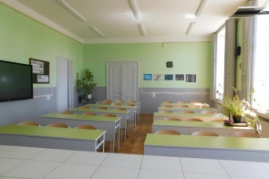 Sala lekcyjna z równo ustawionymi ławkami i krzesłami, tablicą interaktywną na ścianie, dużymi oknami po prawej stronie oraz roślinami doniczkowymi na parapetach.