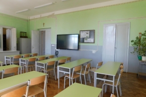 Sala lekcyjna z zielonymi ścianami, ustawionymi w rzędach ławkami i krzesłami, tablicą interaktywną na ścianie, dwoma białymi drzwiami oraz dużą rośliną w doniczce.