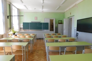 Sala lekcyjna z ustawionymi w rzędach ławkami i krzesłami, klasyczną zieloną tablicą na froncie, tablicą interaktywną z boku oraz dużymi oknami po lewej stronie.