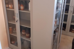 Metalowe szafy z przeszklonymi drzwiami, wewnątrz których znajdują się szklane butelki i pojemniki laboratoryjne, ustawione w pomieszczeniu z jasną podłogą.