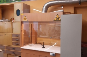 Stanowisko laboratoryjne z drewnianą szafką i dygestorium, wyposażone w zlew, kurki wodne oraz osłonę ochronną, na ścianie widoczne układy okresowe pierwiastków.