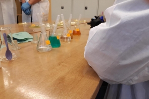 Stół laboratoryjny z kolbami i zlewkami wypełnionymi kolorowymi cieczami, osoby w fartuchach laboratoryjnych stoją w tle.