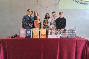 Zdjęcie przedstawia autorkę książek - panią Ninę Majewską-Brown, nauczycielkę języka polskiego - panią Darię Mazur oraz grupę uczniów z oddziału 1TI,  oraz zdjęcia kilku książek autorki 