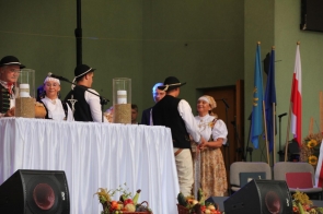 Uroczystość dożynkową odbywającą się na scenie. Na pierwszym planie znajduje się stół przykryty białym obrusem, na którym ustawiono dwa wysokie szklane naczynia wypełnione ziarnem oraz krucyfiks. Na stole widoczny jest także duży bochenek chleba, symbolizujący urodzaj i wdzięczność za plony. Wokół stołu stoi kilka osób ubranych w tradycyjne stroje ludowe. Po lewej stronie znajduje się mężczyzna (wójt Andrzej Kozieł) w kamizelce z haftami i kapeluszu, trzymający chleb. Obok niego stoi kobieta w białej bluzce (starosta Janina Żagan), zdobionej kamizelce i chustce na głowie. Dalej po prawej stronie dwóch mężczyzn ubranych w ludowe kamizelki i kapelusze stoi twarzą do siebie, jakby wymieniając się gestami przyjaźni lub gratulacjami. Za nimi stoi mężczyzna (Maciej Iskrzycki) w garniturze oraz kobieta (Wioleta Iskrzycka) w tradycyjnym stroju z chustą na głowie, która patrzy z uśmiechem. W tle widoczne są flagi Unii Europejskiej, Powiatu Cieszyńskiego i Polski, co nadaje wydarzeniu oficjalny charakter. Cała scena jest starannie udekorowana i przygotowana, oddając ducha tradycji i wspólnoty, które towarzyszą świętu dożynek. Atmosfera na zdjęciu jest uroczysta, pełna szacunku i radości z zakończonych żniw.
