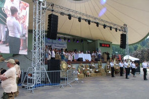 Uroczystość dożynkową odbywającą się na dużej scenie pod zadaszeniem w Amfiteatrze im. Elżbiety Bauer w Brennej. Na scenie znajduje się grupa osób ubranych w tradycyjne stroje ludowe. Na środku sceny stoi stół przykryty białym obrusem, a wokół niego ustawione są wieńce dożynkowe wykonane z kłosów zbóż i ozdobione kwiatami.  Po prawej stronie sceny widać zegar elektroniczny wskazujący godzinę 16:16:09. Na pierwszym planie, przed sceną, znajduje się zespół muzyczny grający na instrumentach dętych, który dodaje muzycznej oprawy całemu wydarzeniu. W tle sceny wisi transparent z napisem "Powiatowo-Gminne Dożynki Ekumeniczne w Brennej", co wskazuje na oficjalny charakter uroczystości oraz współpracę lokalnych wspólnot religijnych.  Po lewej stronie zdjęcia widać duży ekran, na którym wyświetlane są fragmenty ceremonii, ułatwiając widzom obserwację tego, co dzieje się na scenie. Wokół sceny stoi kilka osób, w tym ochrona w beżowych koszulkach, pilnująca porządku. Atmosfera na zdjęciu jest podniosła i pełna tradycji, oddając ducha świętowania zakończenia żniw i dziękczynienia za plony.