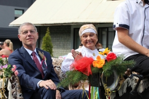 Po lewej stronie siedzi wójt Andrzej Kozieł ubrany elegancko w granatowy garnitur, białą koszulę i bordowy krawat. Na klapie jego marynarki przypięta jest ozdoba z kłosa zboża i różowej wstążki, co nawiązuje do dożynkowego wydarzenia. Obok niego, po prawej stronie, siedzi Janina Żagan Starosta Cieszyński, ubrana w tradycyjny strój ludowy, z białą bluzką zdobioną koronkami i złotą broszką oraz z chustą na głowie. Janina Żagan ma pogodny wyraz twarzy i szeroko się uśmiecha, machając do publiczności. Wóz, na którym siedzą, jest udekorowany zielonymi gałązkami, kolorowymi kwiatami i wstążkami, co dodaje całości radosnego, świątecznego charakteru. W tle widoczny jest budynek, co sugeruje, że zdjęcie zostało zrobione na ulicy w trakcie dożynkowego korowodu. Atmosfera na zdjęciu odzwierciedla ducha świętowania i wspólnoty.