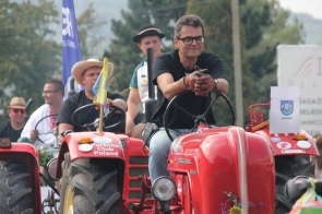 Uczestnicy dożynek jadący na czerwonym traktorze, który jest częścią korowodu w Brennej. Na pierwszym planie siedzi Rajmund Dedio, Burmistrz Skoczowa w czarnej koszulce i okularach, uśmiechający się lekko. Ma na sobie brązowe rękawiczki, na jego nadgarstku jest zegarek. Na przodzie traktora znajduje się logo „Porsche Club Poland”. Za nim, na kolejnym traktorze, siedzi mężczyzna w kapeluszu, trzymający kierownicę oraz inna osoba w tradycyjnym stroju ludowym, co podkreśla różnorodność uczestników i pojazdów w korowodzie. W tle można dostrzec inne osoby i pojazdy, co sugeruje, że korowód jest duży i różnorodny. Na traktorze widoczna jest również flaga oraz kartka z herbem Skoczowa. Zdjęcie ukazuje radosną atmosferę wydarzenia i zaangażowanie lokalnej społeczności w przygotowanie tego tradycyjnego święta.