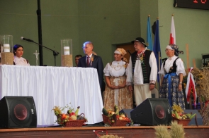 Uroczystość dożynkową na scenie, gdzie grupa osób bierze udział w ceremonii. Na pierwszym planie znajduje się stół przykryty białym obrusem, na którym ustawiono dwa wysokie szklane naczynia wypełnione ziarnem oraz krucyfiks. Stół zdobią także kosze z owocami i warzywami, symbolizujące obfitość plonów i dary ziemi. Po lewej stronie stołu siedzi kobieta ubrana w tradycyjny strój ludowy z białą bluzką i ciemnym gorsetem. Obok stołu, po prawej stronie stoją cztery osoby: elegancko ubrany mężczyzna w garniturze (wójt Andrzej Kozieł) z przypiętą ozdobą dożynkową na klapie marynarki oraz trzy osoby w ludowych strojach. Kobiety (Janina Żagan i Wioleta Iskrzycka) mają na sobie kolorowe fartuchy, białe bluzki i chusty na głowach, a mężczyzna jest ubrany w tradycyjną kamizelkę z haftami i czarny kapelusz. Wszystkie osoby patrzą na stół, co potwierdza, że uczestniczą w oficjalnej ceremonii związanej z dożynkami. W tle widoczne są flagi Unii Europejskiej, Powiatu Cieszyńskiego i Polski, co podkreśla uroczysty charakter wydarzenia. Atmosfera na zdjęciu jest poważna i pełna szacunku dla tradycji, co widać w skupionych twarzach uczestników oraz starannie przygotowanej dekoracji sceny.