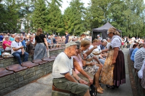 Grupa osób siedzących na ławkach w amfiteatrze podczas dożynek. Uczestnicy wydarzenia, w różnym wieku, są rozlokowani na widowni na świeżym powietrzu, otoczeni drzewami. Wśród zgromadzonych dominują osoby starsze, które siedzą i rozmawiają, niektórzy z nich trzymają w rękach przekąski lub napoje. Na pierwszym planie po prawej stronie widać starostę Janinę Żagan ubraną w tradycyjny strój ludowy: białą bluzkę z koronką, wzorzysty fartuch i chustę na głowie. Starosta częstuje uczestników dożynkowego święta chlebem, który trzyma w koszyku. Uśmiecha się i rozmawia z osobami siedzącymi na ławkach, co wskazuje na serdeczną atmosferę panującą podczas tego wydarzenia. W tle widoczne są inne osoby, które również cieszą się uroczystością, rozmawiają lub obserwują to, co dzieje się na scenie. Otaczające amfiteatr drzewa dodają przytulności i tworzą naturalną, zieloną scenerię. Zdjęcie oddaje atmosferę wspólnoty i tradycji, pokazując, jak lokalna społeczność wspólnie świętuje zakończenie żniw i obfitość plonów.