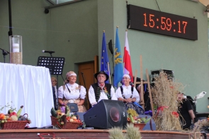 Na zdjęciu widoczna jest scena podczas dożynek, na której znajdują się osoby ubrane w tradycyjne stroje ludowe. Są nimi od lewej strony: Janina Żagan, starosta, Andrzej Kozieł, wójt oraz Wioleta Iskrzycka, gadzina dożynkowa. Na pierwszym planie po lewej stronie stoi stół przykryty białym obrusem, na którym ustawiono dużą szklaną wazę z ziarnem. Przed stołem widać kosze wypełnione owocami, warzywami i zbożem, które symbolizują urodzaj i dary ziemi. Kobiety mają na sobie białe bluzki z koronkowymi zdobieniami i kolorowe chusty na głowach, a mężczyzna jest ubrany w kamizelkę zdobioną tradycyjnymi wzorami oraz kapelusz. Wszyscy mają radosne wyrazy twarzy, co sugeruje, że są zadowoleni z udziału w uroczystości. W tle, za nimi, widoczne są flagi: Unii Europejskiej, powiatu cieszyńskiego i Polski, co podkreśla oficjalny charakter wydarzenia. Na ścianie wisi duży zegar elektroniczny, pokazujący godzinę 15:59:17. Obok sceny widać dekoracje z suszonych zbóż i słomy, co dodatkowo wzmacnia tradycyjny i dożynkowy charakter uroczystości. Atmosfera na zdjęciu jest uroczysta i pełna dumy z dziedzictwa kulturowego oraz rolniczych tradycji.