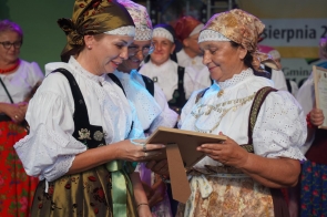 Na zdjęciu znajduje się grupa kobiet ubranych w tradycyjne stroje ludowe, które są bogato zdobione. Kobiety mają na sobie białe bluzki z koronkowymi kołnierzami oraz fartuchy w różnorodne wzory. Niektóre z nich mają na głowach chusty. Dwie kobiety (od prawej strony Janina Żagan Starosta) rozmawiają ze sobą. Kobieta po lewej stronie, w ciemnym gorsecie i białej bluzce, trzyma w rękach dyplom i z uśmiechem rozmawia z drugą kobietą, która również przygląda się dyplomowi. Starosta, ubrana jest w jasny, kwiecisty fartuch, białą bluzkę i jasny gorset. Na szyi ma łańcuszek, a na głowie jasną chustę. W tle widać inne kobiety, również ubrane w tradycyjne stroje, które stoją w grupie i trzymają w rękach dyplomy oraz inne nagrody. Atmosfera na zdjęciu jest radosna. 