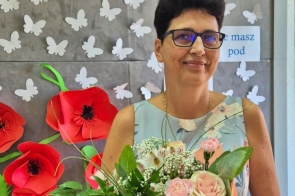 Bożena Kania-Sitek, Dyrektorka Zespołu Szkół Ekonomiczno-Gastronomicznych w Cieszyna ma założoną na sobie sukienkę w kwiaty, nosi okulary. W ręce trzyma bukiet różowych kwiatów, który otrzymała od władz Powiatu. Stoi na tle dekoracji z papierowymi motylami i kwiatami, z napisem „ZSEG w Cieszynie”. 
