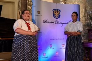 Od lewej Anna Musioł oraz Izabela Orawska ubrane w tradycyjne stroje ludowe. W tle widoczna podświetlona na niebiesko ścianka z herbem Powiatu i napisem Powiat Cieszyński.