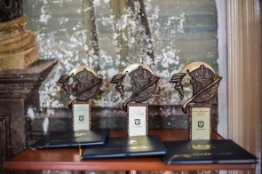 Na brązowym stoliku stoją 3 statuetki dla laureatów oraz 3 listy gratulacyjne, znajdujące się w granatowych, eleganckich teczkach.