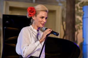 Aneta Legierska-Bujok przemawia przez bezprzewodowy mikrofon. Ubrana jest w białą, elegancką koszulę. Włosy ma spięte w kok, nad uchem ma przypięty czerwony kwiat. Kobieta jest uśmiechnięta.