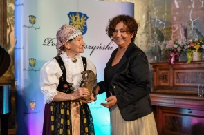 Na pierwszym planie, od lewej Helena Wróbel, ubrana w tradycyjny strój ludowy, trzyma w ręku statuetkę. Tuż obok niej Stoi Janina Żagan Starosta Cieszyński. Obie Panie są uśmiechnięte. W tle widoczna jest niebieska ścianka z herbem i napisem Powiat Cieszyński