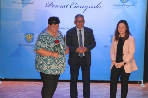 Trzy osoby stoją na scenie podczas oficjalnej uroczystości. Osoba po lewej trzyma różę i dyplom. W tle widoczny jest niebieski baner z herbem Powiatu Cieszyńskiego.