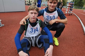 Na zdjęciu dwóch młodych sportowców w strojach biegowych. Jeden siedzi na tartanowej bieżni, drugi klęczy obok, obejmując go ramieniem. Obaj mają na sobie numery startowe. W tle widoczne są inne osoby i elementy stadionu lekkoatletycznego. Atmosfera sportowej rywalizacji.