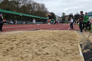 Sportowiec wykonuje skok w dal na piaszczystą strefę zeskoku. W tle widać trybuny z zadaszeniem, innych uczestników zawodów oraz osoby obserwujące rywalizację. Pogoda jest pochmurna, ale atmosfera wydaje się skupiona i pełna energii.