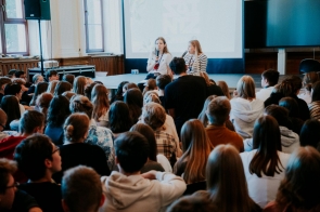 W sali z wysokimi oknami i jasnym wnętrzem zgromadziła się grupa młodzieży, siedząca na podłodze i krzesłach, uważnie słuchająca dwóch kobiet, które prowadzą spotkanie z mikrofonami w dłoniach. W tle znajduje się ekran z prezentacją. Atmosfera jest skupiona i edukacyjna.