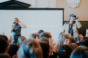 Na zdjęciu widzimy dwójkę artystów na scenie podczas występu. Kobieta w dżinsowym stroju oraz mężczyzna w beżowej kurtce i niebieskiej czapce trzymają mikrofony, wykonując utwór. W tle widać wyświetlany napis, a przed sceną tłum ludzi z uniesionymi rękami, tworzących energetyczną atmosferę koncertu.