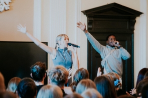 Na zdjęciu widzimy dwójkę osób na scenie podczas występu. Kobieta w stroju jeansowym i mężczyzna w szarej bluzie trzymają mikrofony i gestykulują z uniesionymi rękami, zachęcając publiczność do wspólnej zabawy. Przed nimi znajduje się tłum osób z uniesionymi rękami, tworzących żywą atmosferę koncertu.